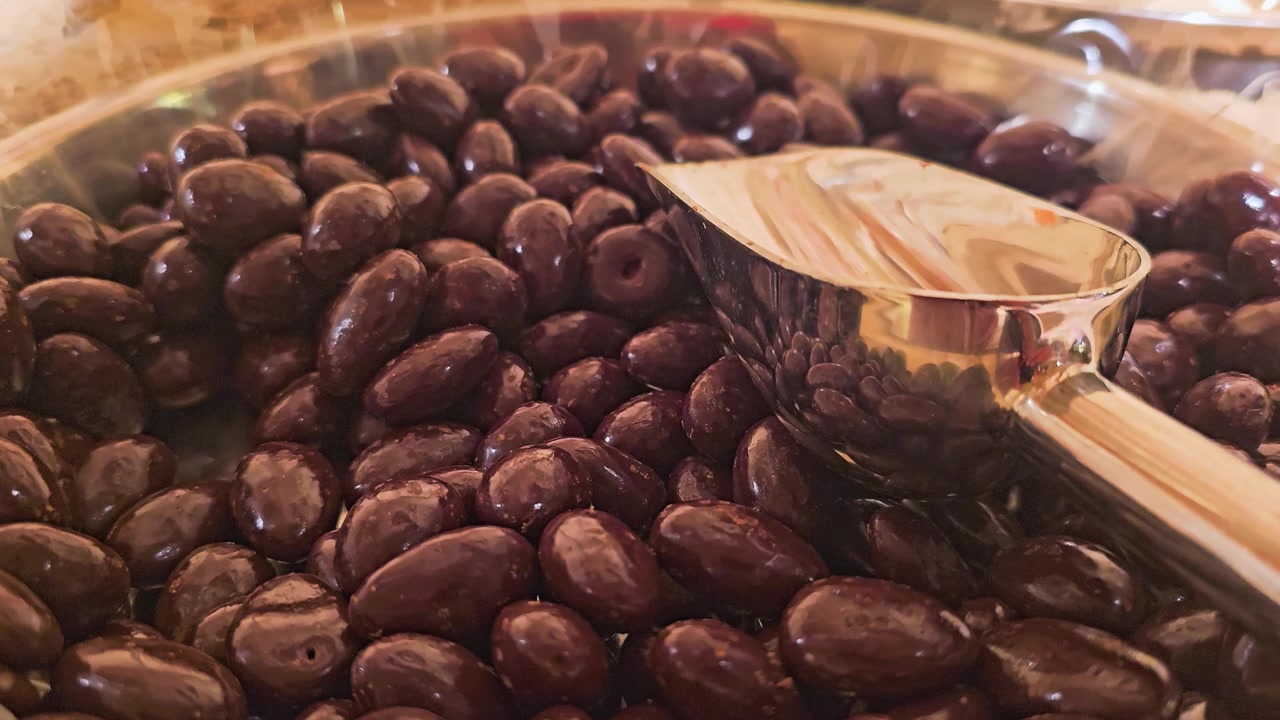 Gourmet confectionery display with dark chocolate almonds and stainless scoop in a self serve sweets bin, showing glossy surfaces, rich brown tones, and cozy shop lighting.