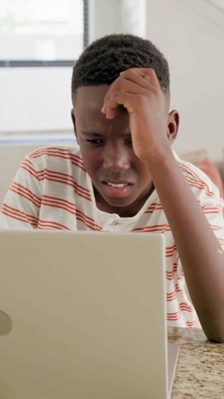 Vertical video: Reacting teen boy in striped shirt reading laptop post at home, typing to respond