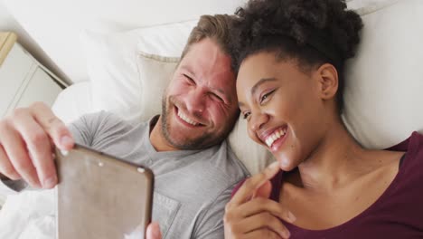 video of happy diverse couple using tablet in bed and talking