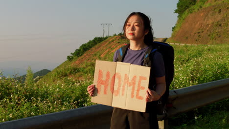 Junge-Asiatische-Anhalterin,-Die-Mit-Einem-Schild,-Auf-Dem-Ihr-Reiseziel-Angegeben-Ist,-Nach-Hause-Gehen-Möchte