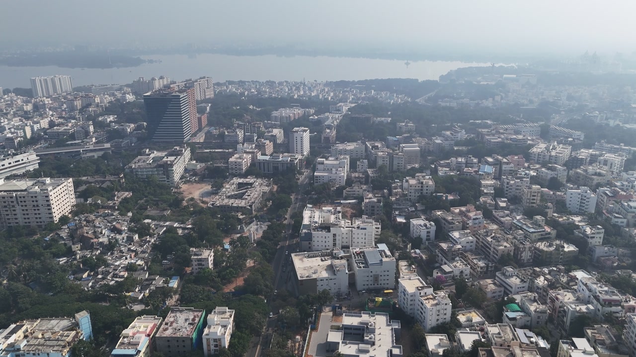 Daytime aerial view near Hussain Sagar, featuring dense residential blocks, commercial structures, and green spaces across a growing city environment.