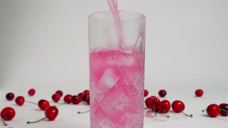 static medium shot of a glass filled with ice cold and clear ice cubes while a cherry drink is poured with red cherries in the background in slow motion