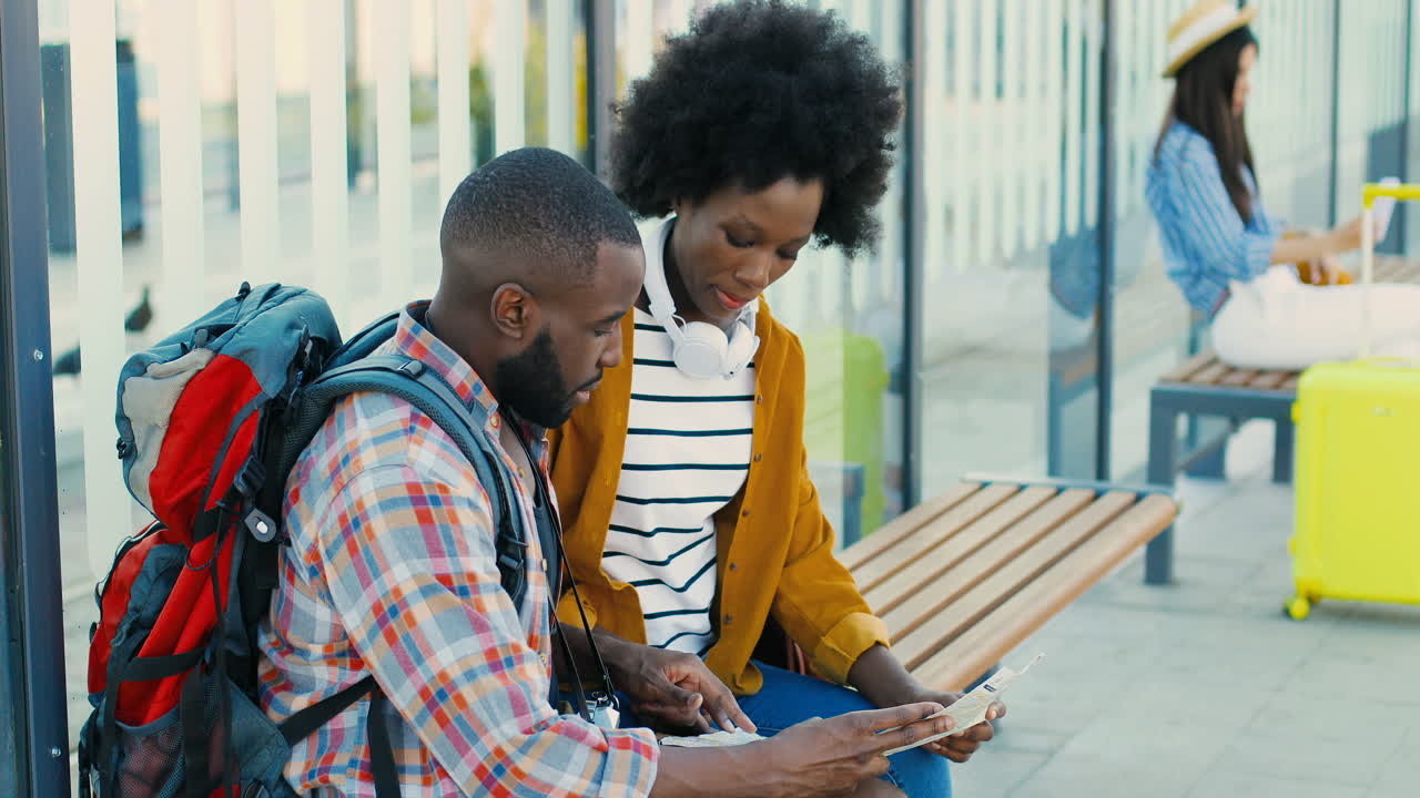 African american young happy man and woman travellers sitting at bus stop talking and watching a map to plan a route