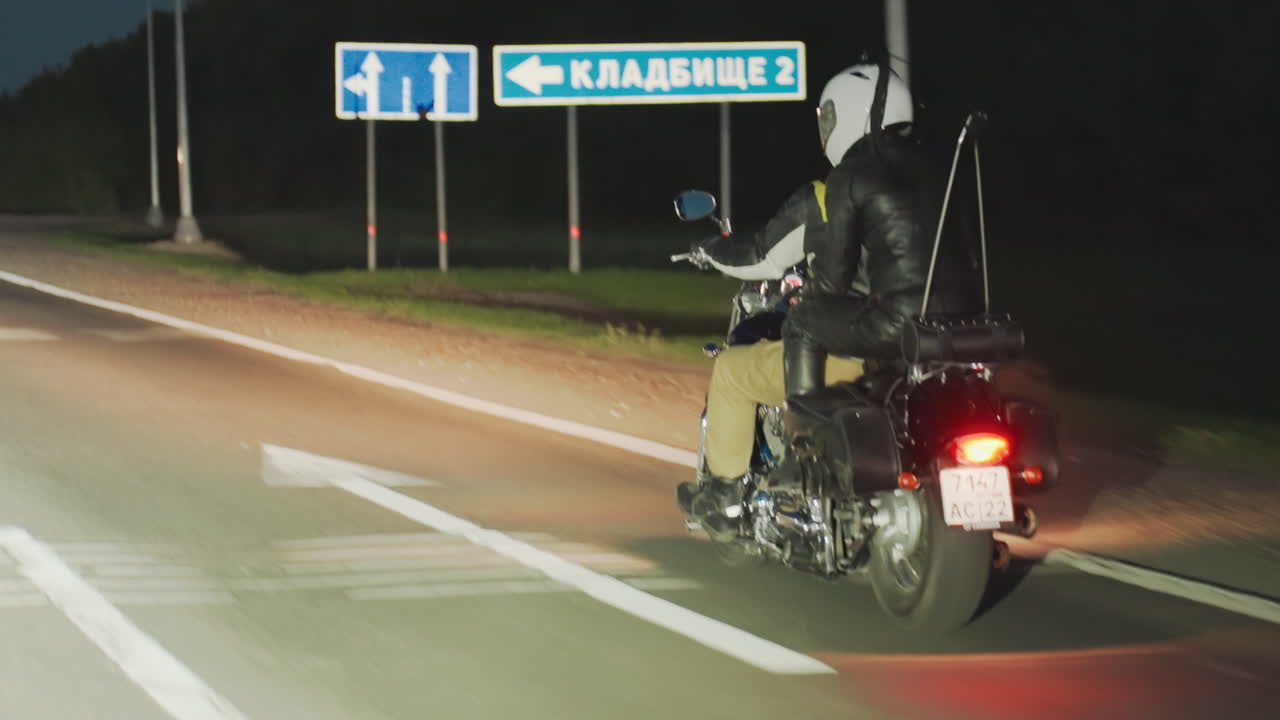 Couple on motorcycle ride at night along highway, rear light glowing as they pass illuminated signpost ahead, wearing helmets and leather jackets while traveling under dark evening sky