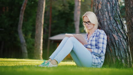 a blonde young woman in glasses reads a book in the park sits near a tree beautiful light before sun