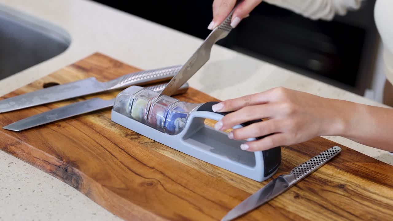 Hands sharpening a knife using a manual sharpener on a wooden board in a well-lit kitchen setting