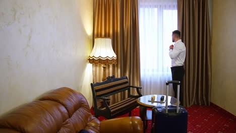 young business man in a hotel room. man in elegant clothing standing near the window. traveler arrived to a hotel with suitcase. business journey.