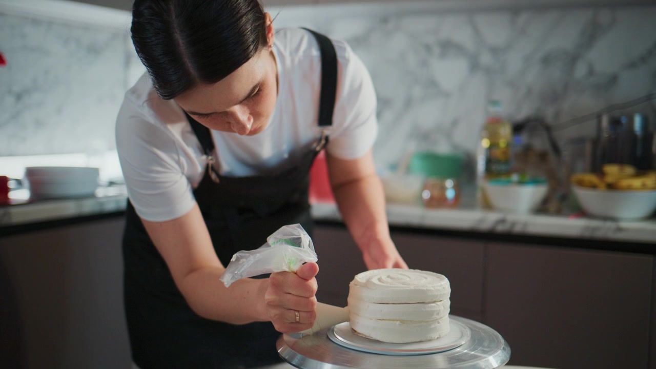 Fokusert kvinnelig baker sprøyter forsiktig glasur på en etasjekake. Hun lener seg fremover med konsentrasjon, hendene er stødige og presise. Kjøkkenet har rene, moderne overflater med myk belysning.