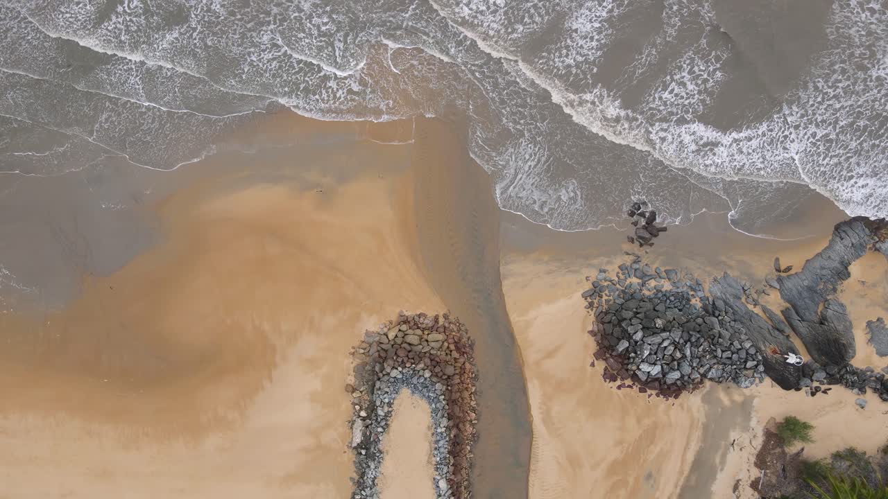 video aéreo de drones mirando hacia abajo en una playa desierta con olas rompiendo hasta un arroyo que fluye hacia el océano