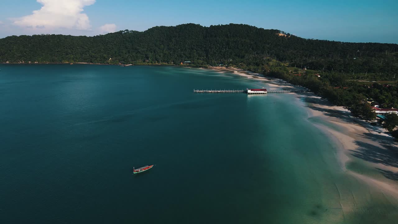 Beautiful clear blue sea with a long stretch of jetty built in the calm sea with large tourist resorts in the background between green nature. Wide drone panning shot