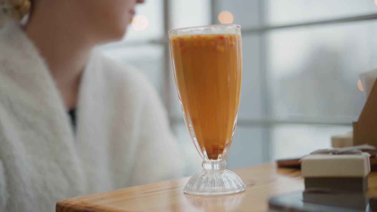 Un bicchiere alto con stelo pieno di tè freddo ambrato si trova su un tavolo di legno accanto a una finestra luminosa, cubetti di ghiaccio e condensa mostrano un freddo rinfresco, mentre una persona sfocata in accappatoio sta nelle vicinanze in