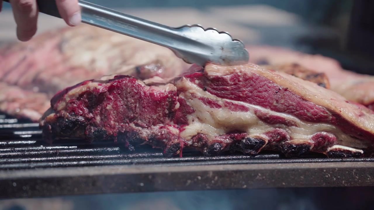 Traditional Argentine asado de tira grilling over a wood fire while tongs turn lamb ribs in the background surrounded by smoke and intense heat. Authentic outdoor barbecue cooking scene.