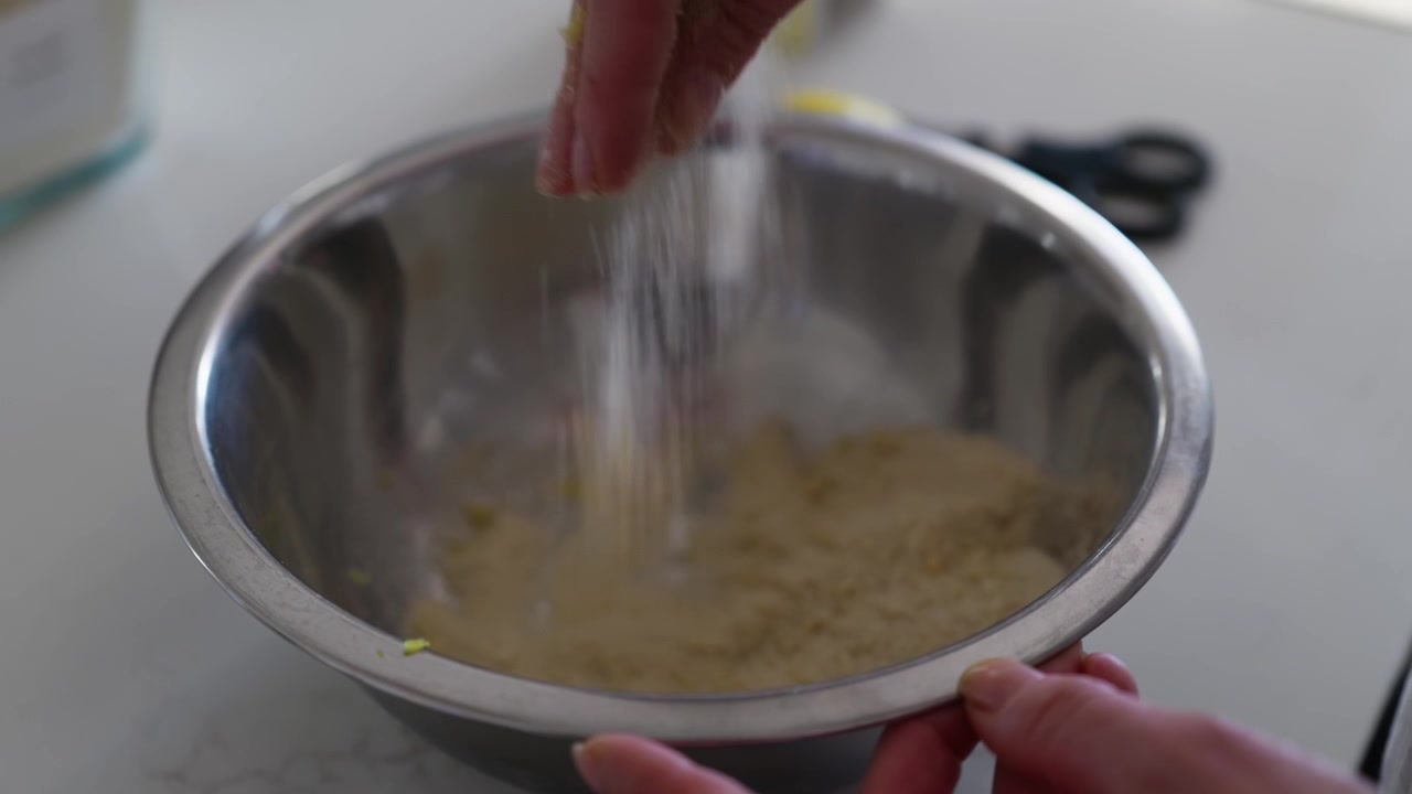 Primo piano di mani che sbriciolano scorza di limone fresca nella farina in una ciotola. Gli ingredienti vengono combinati per la cottura, mettendo in risalto la consistenza, la preparazione e la cucina casalinga