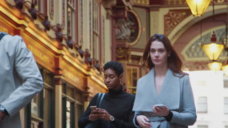 Millennial-business-people-walking-towards-camera-in-London’s-historical-Leadenhall-covered-street-market,-checking-the-time-and-using-their-phones,-low-angle