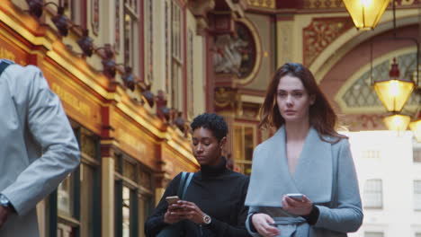 millennial business people walking towards camera in london’s historical leadenhall covered street market, checking the time and using their phones, low angle