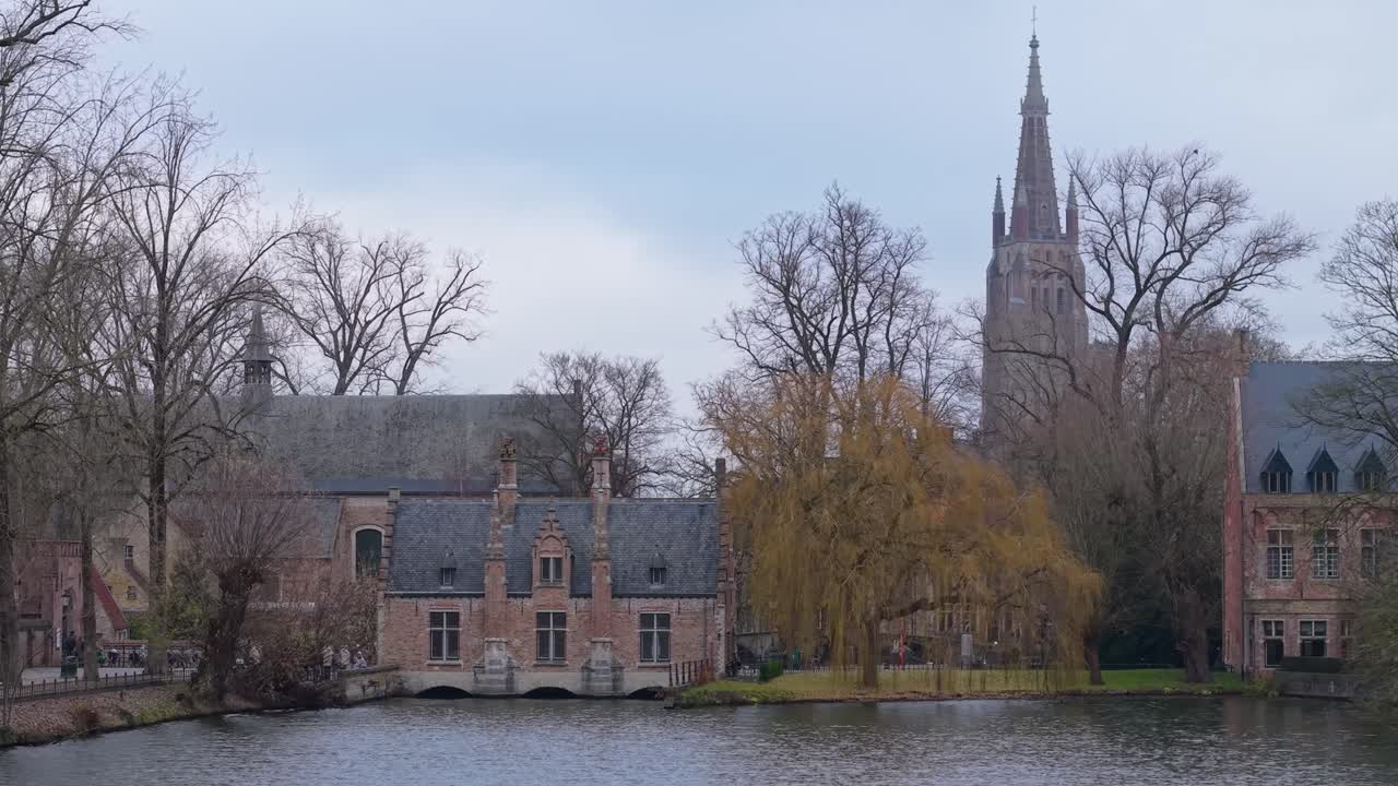 Filmato laterale del Sashuis di Bruges con alberi circostanti, un lago e il campanile sullo sfondo, che mette in risalto lo storico e suggestivo paesaggio urbano.