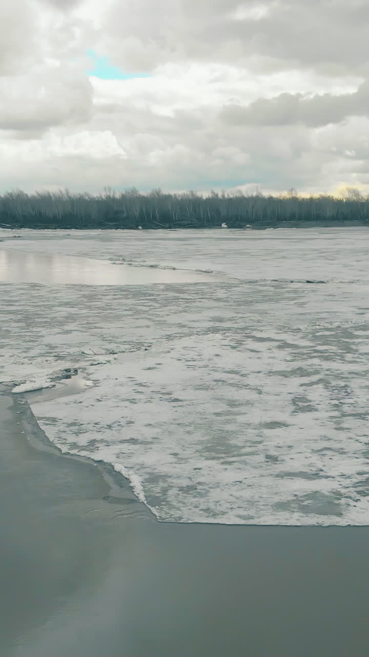 deep frozen river against bank with dense forest on horizon at grey bridge under cloudy sky upper view