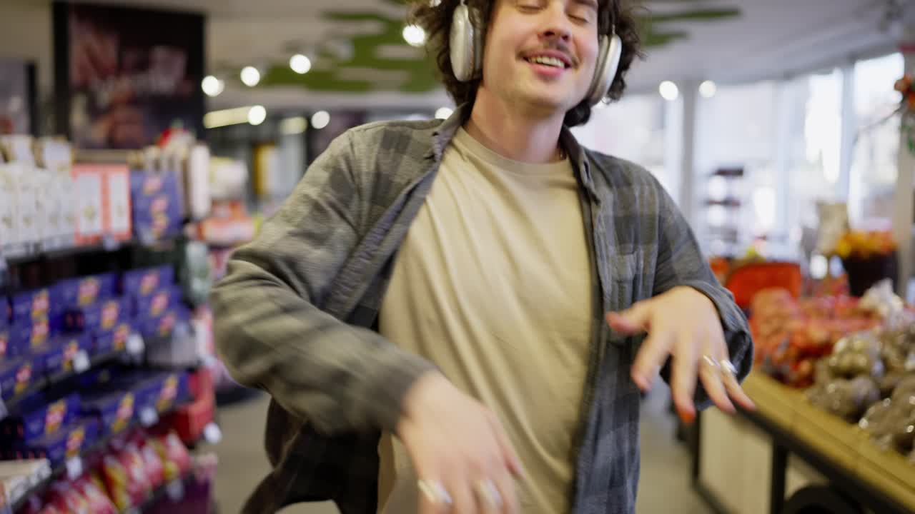 Joyful brunette guy in a checkered shirt with white headphones listens to music walks and dances intensely in a grocery store while shopping
