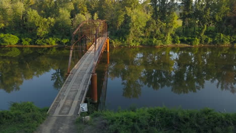 toma aérea de derecha a izquierda de un viejo puente de hierro sobre un río rural bordeado de árboles en la cálida luz de la tarde