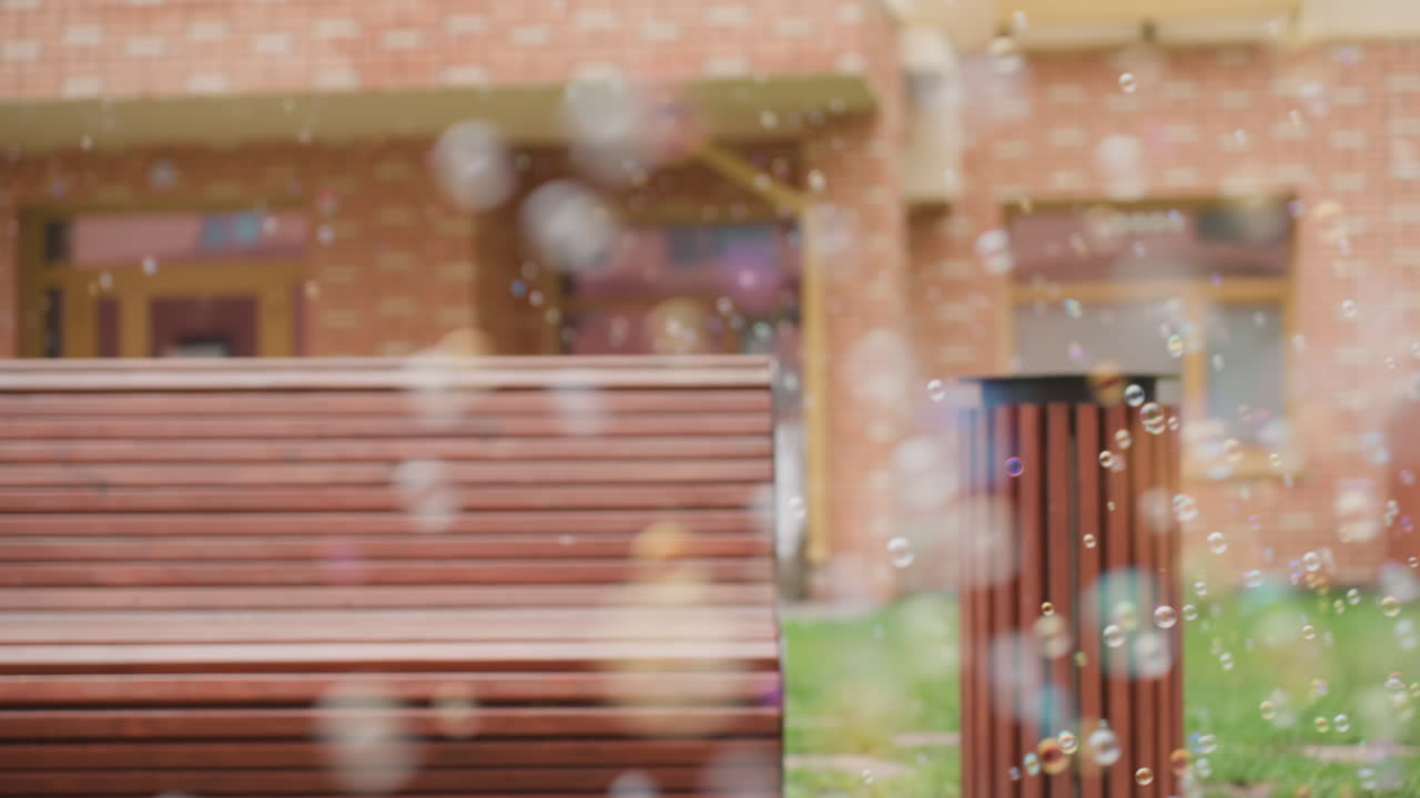Close up of bubbles floating across courtyard in front of bench and brick building, gentle drift over grass, soft blur background adds sparkle and calm mood during quiet outdoor moment