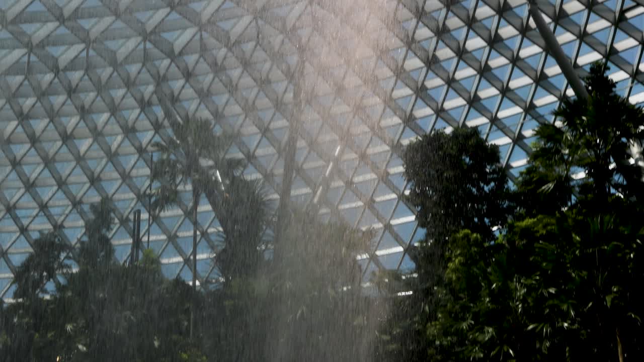 A dramatic indoor waterfall pours from a glass-domed ceiling into a verdant garden, with natural daylight illuminating geometric patterns and tropical plants. Static camera, vertical framing
