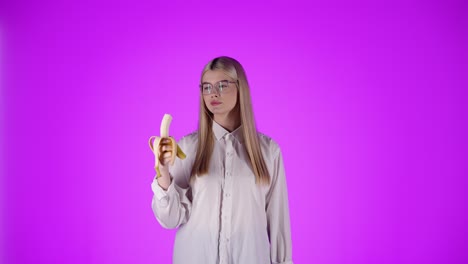 Junge-Schöne-Kaukasische-Blonde-Frau-Mit-Brille-Isst-Eine-Banane-Tief-In-Ihrem-Mund-Studio-Professionellen-Hintergrund-Oberkörperaufnahme