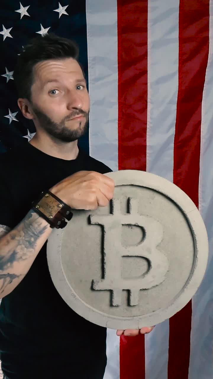 Tattooed man with short hair and beard raising up a large concrete and gray colored Bitcoin cryptocurrency sculpture art and nodding with approval in front of a vertical American US flag, patriot
