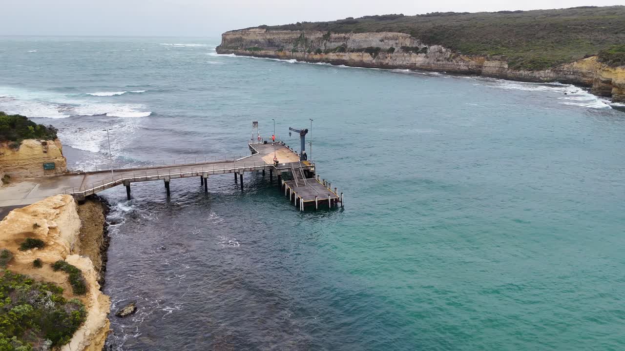 Drone footage captures Port Campbell's pier and rugged coastline, showcasing turquoise waters and dramatic cliffs under overcast skies