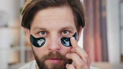 concepto de cuidado facial donde simpático sonriente cariñoso joven barbudo tocando parches de hidrogel negro debajo de los ojos y mirando a la cámara