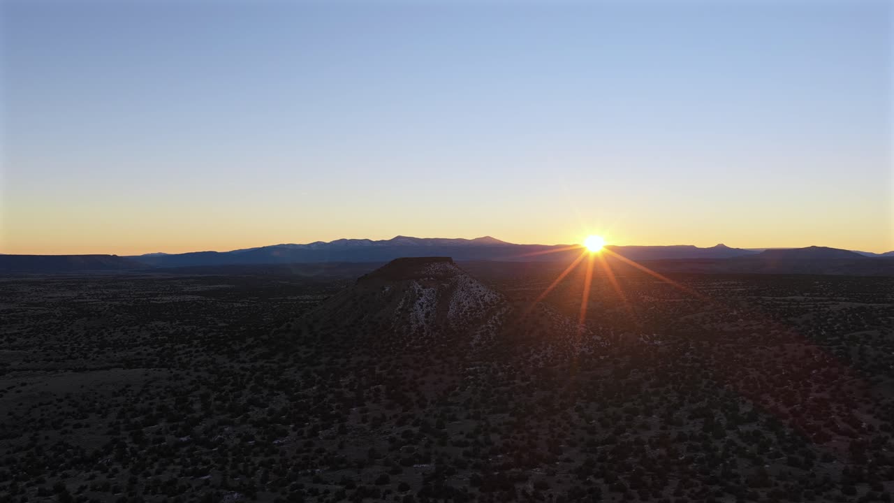 Bredt dronebilde av en vulkansk slaggkjegle som stiger opp fra høye ørkensletter. Den lave vinkelen på solen skaper en dramatisk silhuett, og fremhever den robuste teksturen i landskapet i New Mexico.