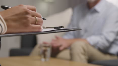 close up of female therapist or counsellor taking notes in conversation with mature man in office