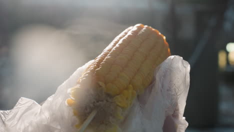 close up of partially eaten corn being turned with steam rising, bokeh lights in background, soft lighting illuminating kernels, fresh food, plastic wrap still around corn