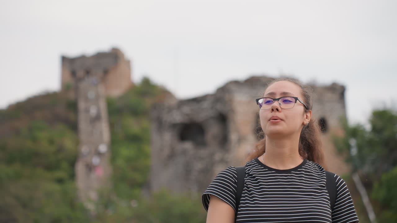 Jovem mulher em ruínas olhando para cima, protegendo os olhos, caminhante casual explorando uma antiga torre de pedra em uma encosta verde, óculos, céu nublado, expressão contemplativa, caminhada lenta, clima de viagem.