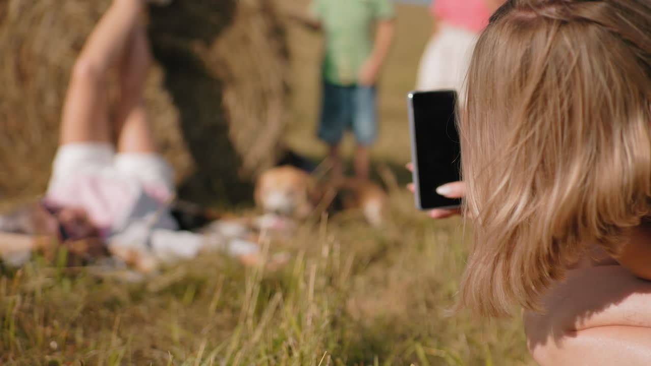 близкий задний вид женщины, фотографирующей человека, лежащего на сене с поднятыми ногами, в окружении пышного зеленого поля, размытый вид двух детей, играющих в фоновом режиме