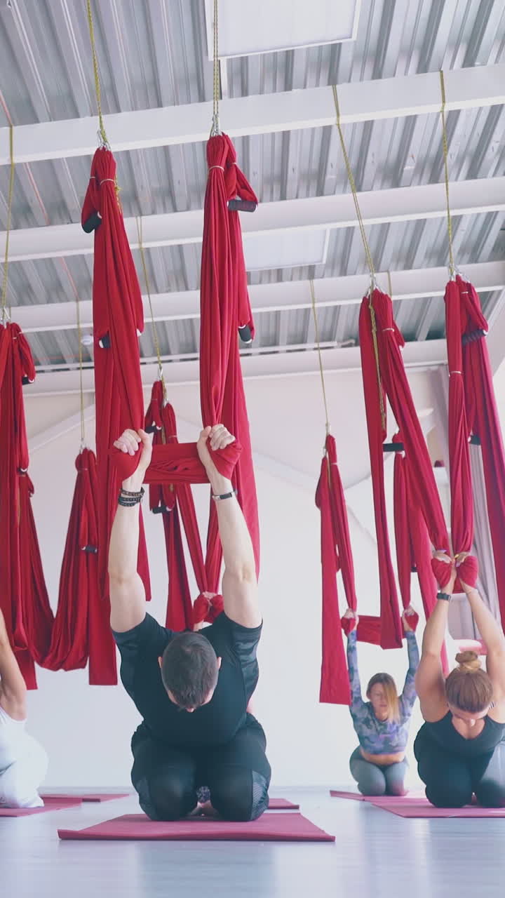 advanced group stretches leg muscles holding red hammocks after hard oriental fly yoga workout in white room
