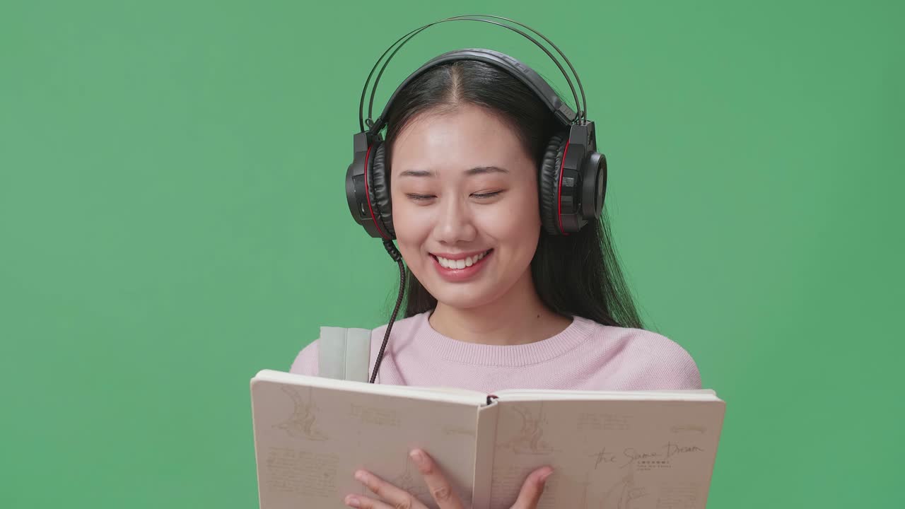 아시아 여성 학생이 책을 읽고 녹색 스크린 스튜디오에서 헤드폰으로 음악을 듣고 있습니다.