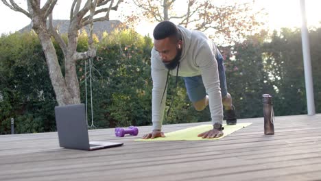 Hombre-Afroamericano-Enfocado-En-Entrenamiento-Físico-Haciendo-Flexiones-En-Cubierta-En-Un-Jardín-Soleado,-Cámara-Lenta