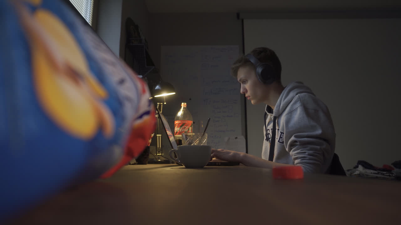 joven tomando un refrigerio, bebiendo refrescos e impacientemente tratando de llegar a una comida chatarra envuelta al otro lado de la mesa mientras hace algo en su computadora portátil en casa