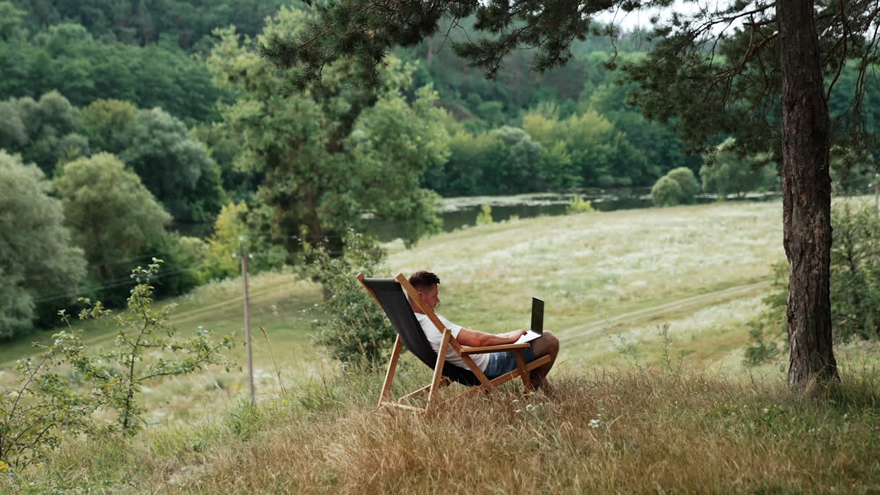 Freelancer works in a beautiful green place away from the city. Man sits in a folding chair with laptop on his knees.