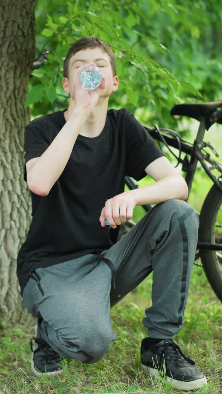 두 어린 형제는 나무 에 앉아서 물병에서 물을 마시며 자전거 타고 휴식을 취하고 있으며, 두 자전거는 평화로운 숲의 울창한 초록색으로 둘러싸여 근처에 주차되어 있습니다.