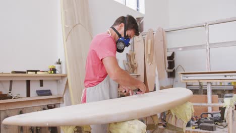 Fabricante-De-Tablas-De-Surf-Masculino-Caucásico-Con-Una-Máscara-Facial-Y-Preparándose-Para-Pulir-Una-Tabla-De-Surf