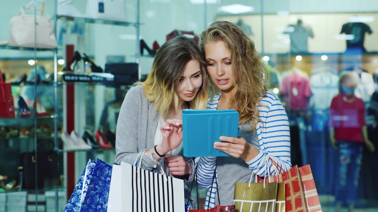 태블릿에서 사용하는 쇼핑백을 들고 있는 두 여자 친구는 유리창 뒤에 서 있습니다.