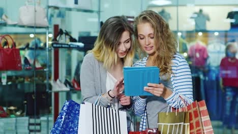Two-Woman-Friends-With-Shopping-Bags-Used-By-The-Tablet-They-Stand-On-A-Background-Of-Glass-Windows-