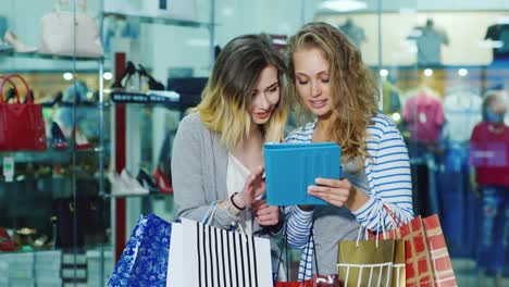 two woman friends with shopping bags used by the tablet they stand on a background of glass windows