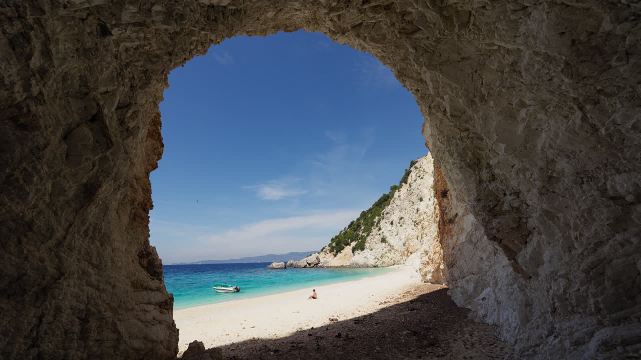 Wide cave view frames man sitting and pondering on Platinamos Beach, Ithaca Island