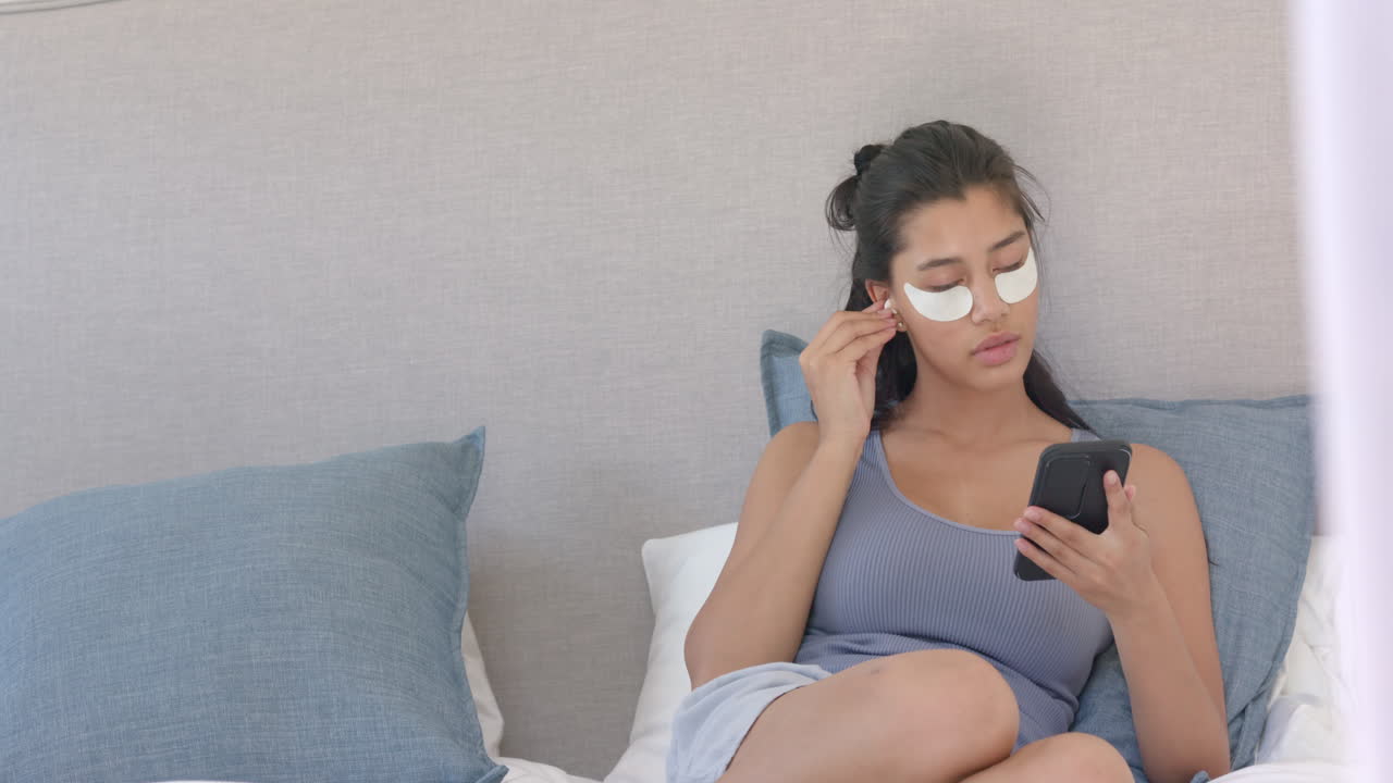 Biracial teenage girl wearing under eye masks sitting bed using smartphone, copy space, slow motion