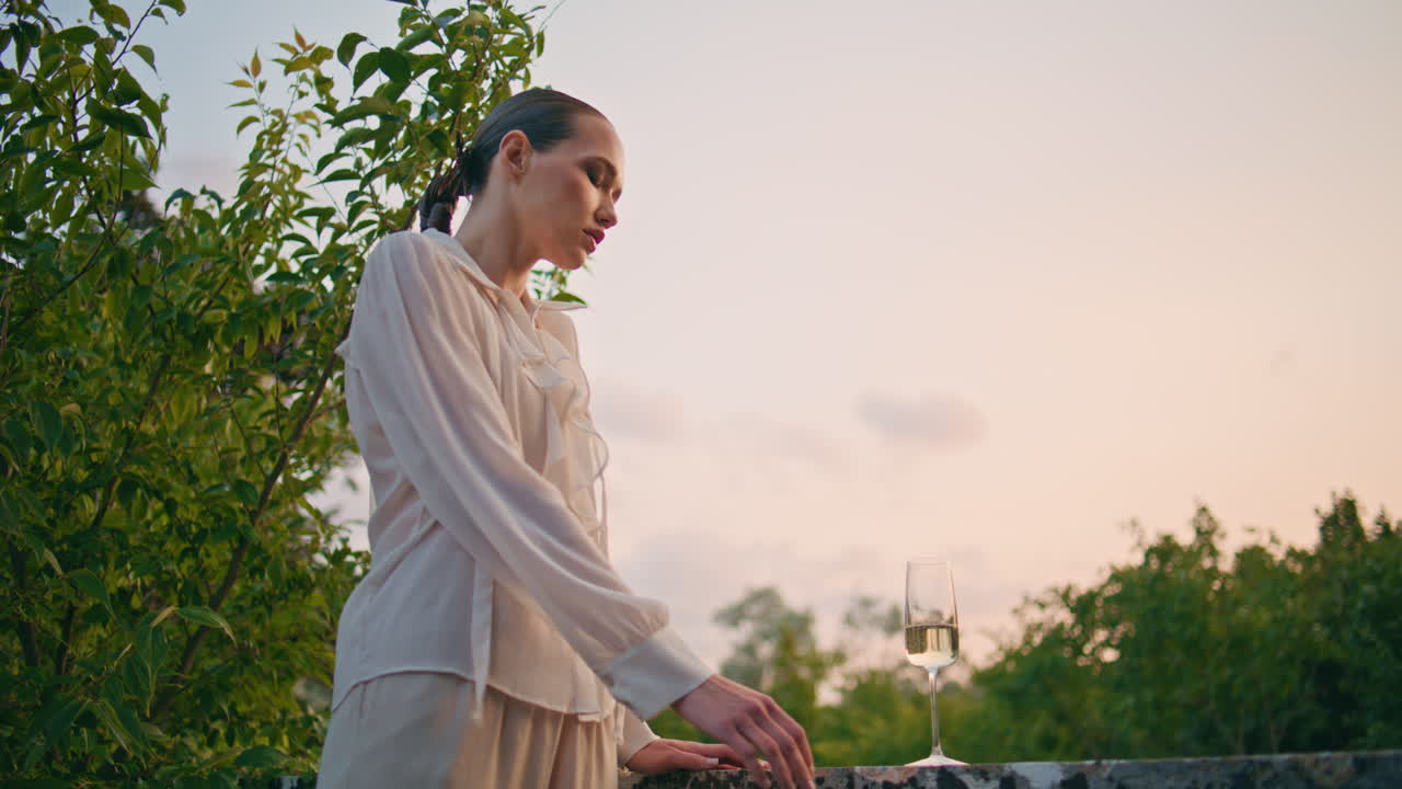 와인 컵과 함께 테라스에서 저녁 아름다움을 찾는 조용한 여성. 발코니에 여인