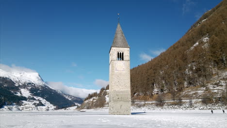Cinematic-aerial-orbit-view-of-sunken-bell-tower-emerging-from-frozen-Lake-Resia-with-snowy-mountains-and-children-playing-in-the-background