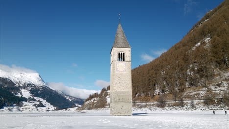 cinematic aerial orbit view of sunken bell tower emerging from frozen lake resia with snowy mountains and children playing in the background