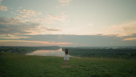 a woman dressed in a white dress and hat stands thoughtfully in a grassy field overlooking a tranquil lake at sunset. she gazes into the distance, creating a serene and reflective scene